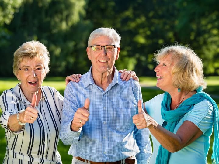 Trzy osoby w letnich ubraniach, z uśmiechem unoszą kciuki. Zielone, rozmyte tło parku. Jasne kolory dominują na zdjęciu.