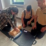 506221791_736597095382481_5354825506364115191_n Na zdjęciu osoba wykonuje resuscytację na fantomie, dwie inne osoby patrzą. Tło to jasny pokój z oknem. Kolory stonowane, dominują beże i brązy.