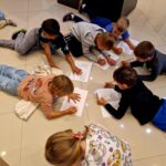 Grupa dzieci leży na podłodze, pisząc na kartkach papieru. Podłoga jest jasna, a dzieci są ubrane kolorowo. Tło jest neutralne, bez widocznych mebli.