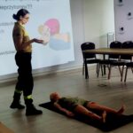 506261389_736596725382518_480759941404807301_n Kobieta w mundurze uczy pierwszej pomocy. Na podłodze leży manekin. W tle rzutnik z instrukcją resuscytacji.