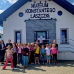 Grupa dzieci i dorosłych stoi przed białym budynkiem z napisem Muzeum Konstantego Laszczki. Budynek ma szare elementy i niebieski dach. W tle widać biało-czerwony dach innego budynku.