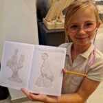 Dziewczynka w okularach trzyma zeszyt z rysunkami ołówkiem. Ma jasne włosy i fioletowy warkoczyk. Tło jest jasne, muzealne z rzeźbami.