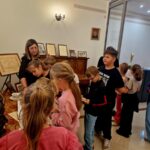 Na zdjęciu grupa dzieci w muzeum zwiedza ekspozycję z przewodniczką. Dzieci są skupione wokół stołu z eksponatami. Tło stanowią obrazy i certyfikaty na ścianach.