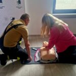 506316054_736596835382507_1998891494440368503_n Na zdjęciu dwie osoby trenują resuscytację na manekinie w sali. Mężczyzna w stroju strażaka, kobieta w różowej bluzce. Tło białe z logo Miasto i Gmina Dobre.