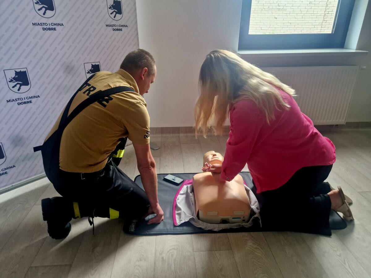 Na zdjęciu dwie osoby trenują resuscytację na manekinie w sali. Mężczyzna w stroju strażaka, kobieta w różowej bluzce. Tło białe z logo Miasto i Gmina Dobre.