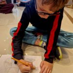 Chłopiec rysuje na siatce w zeszycie, siedząc na podłodze w salonie. Ubrany jest w czarną bluzę z czerwonymi paskami. W tle widać osoby siedzące na podłodze.