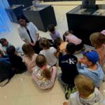 Grupa dzieci siedzi na jasnej podłodze wokół kobiety w białej koszuli. Tło stanowią ciemne gabloty muzealne. Dzieci są skupione i różnorodnie ubrane.