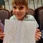 Dziewczynka trzyma szkicownik z rysunkami na kratkowanym papierze. Tło zdobią czarno-białe fotografie na stojakach. Ubrana jest w jasną bluzkę, a jej włosy są jasnobrązowe.