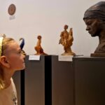 Dziewczynka z opaską patrzy na rzeźby na postumentach. Obok niej popiersie kobiety w chustce. Tło jest jasne z medalionami.