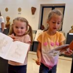 Dwie dziewczynki w muzeum z rysunkami postaci w notatnikach. Tło z popiersiami sztuki na ścianie. Jasne, neutralne wnętrze.