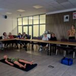 506404420_736597978715726_1845604328087484713_n Grupa siedzi wokół stołu w sali konferencyjnej. Na środku manekin na czarnej macie. Kobieta prowadzi prezentację przy tablicy.