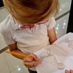 Dziewczynka w okularach rysuje ołówkiem w kratkowanym zeszycie. Ma na sobie białą koszulkę. W tle jasna podłoga.