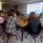 506707865_736597945382396_4500136536215616689_n Grupa ludzi siedzi przy dużym stole i patrzy na ekran z prezentacją w jasnej sali. Na ekranie widoczny jest tekst o powiadamianiu służb ratunkowych. Kolory to głównie biel, brąz i szarość.