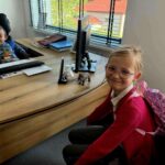 Dwoje dzieci w biurze przy drewnianym biurku. Chłopiec siedzi na fotelu, dziewczynka w różowym plecaku uśmiecha się. W tle okno z żaluzjami.