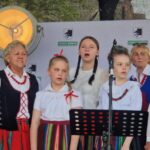 6Pr_i3j1 Grupa osób w strojach ludowych śpiewa na scenie. Tło z logo gminy i dużym reflektorem. Przeważają kolory czerwony, biały i zielony.