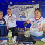 8jIBNaYt Trzy kobiety w ludowych strojach stoją przy stoisku z niebieskim tłem. Jedna z nich gotuje w garnku. Widoczny jest baner Koła Gospodyń Wiejskich.