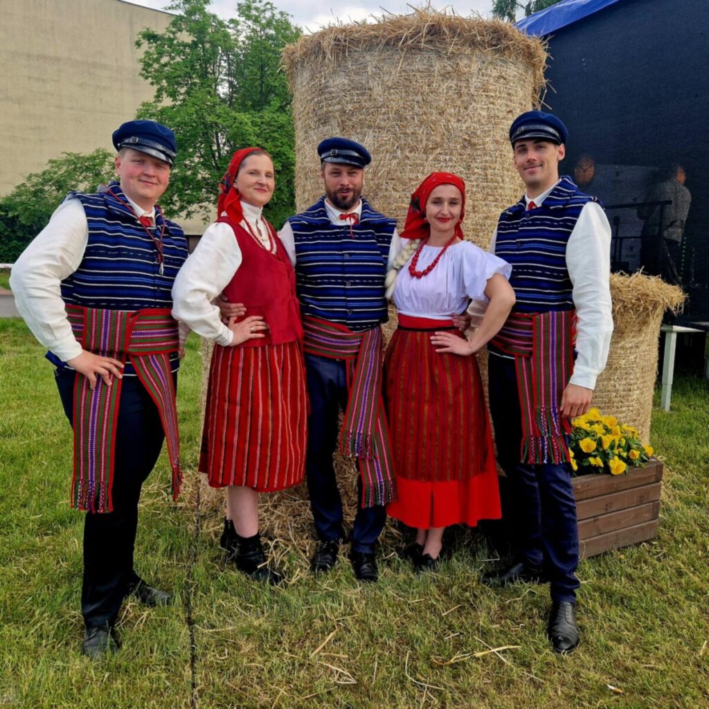 Grupa osób w tradycyjnych strojach ludowych przed belą siana. Mają kolorowe paski i czerwone akcenty. Tło zielone z drzewami.