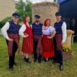 9i3IvTfN Grupa osób w tradycyjnych strojach ludowych przed belą siana. Mają kolorowe paski i czerwone akcenty. Tło zielone z drzewami.