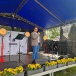 A_3KD2Fu Na scenie pod niebieskim namiotem stoi osoba w jasnej marynarce i dżinsach, przemawiając do mikrofonu. W tle widoczne są reflektory i baner z napisem GMINA DOBRE. Przed sceną znajdują się żółte kwiaty.