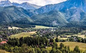 Wiejski krajobraz z widokiem na zielone doliny i góry w tle. W oddali widać małą wioskę i bujną roślinność. Niebo jest jasne, z niewielkimi chmurami.