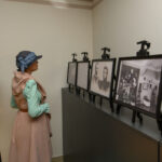 Kobieta w kapeluszu ogląda czarno-białe zdjęcia w galerii. Ubrana w pastelową zieleń, przy jasnej ścianie. Elegancki wystrój.