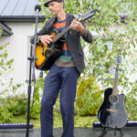 Mężczyzna gra na gitarze na scenie na tle drzew. Ubrany w czapkę, kolorowy sweter i marynarkę. Obok stoi druga gitara.