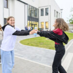 Dwie osoby tańczą przed nowoczesnym białym budynkiem. Dziewczyna w białej bluzie, druga w czerwonej koszulce. W tle zieleń i chodnik.