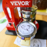 Maszyna do produkcji odznak z czerwonym uchwytem i logo VEVOR. Na metalowej części odznaka z wizerunkiem osoby. Drewniany stół w tle.