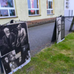 Na zdjęciu widać wystawę plenerową z czarno-białymi fotografiami na tle ściany budynku. Fotografie są ustawione wzdłuż chodnika, ozdobione światełkami. Na trawie obok ustawione są tablice z opisami.