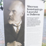 Portret mężczyzny na planszy informacyjnej z tekstem o Muzeum Konstantego Laszczki w Dobrem. Obraz czarno-biały; tło jasne. Na dole widoczne logotypy instytucji.