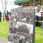 Plakat z czarno-białymi fotografiami historycznymi stoi na zielonej trawie. W tle namiot z lampkami i ludzie. Obok drzewo.