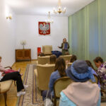 Sala z grupą słuchaczy, kobieta prowadzi wykład. Jasne ściany, żyrandole, zielone zasłony. Obraz orła na ścianie.