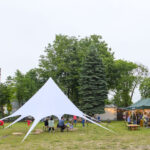 Na zdjęciu widać zielony teren z białym namiotem i ludźmi przy stołach. W tle drzewa i budki z lampkami. Atmosfera jest relaksacyjna.