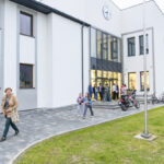 Nowoczesny biały budynek z zegarem. Ludzie wchodzą i wychodzą, otoczeni zieloną trawą. Przed wejściem stoją motocykle i rowery.