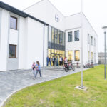 Budynek z białą elewacją i dużymi oknami, przed nim ludzie, rowery i motocykle. Tło z drzewami, zielona trawa. Zegar na ścianie.