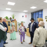 Grupa osób rozmawia w jasnym biurze z białymi ścianami. Jedna osoba ma na sobie zieloną suknię i kapelusz. W tle są rośliny i szafy z drewna.