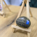 Kamień z napisem na małym drewnianym sztalugowym stojaku. Tło jest rozmyte, drewniany stół w jasnym odcieniu. Na kamieniu widoczna jest niebieska naklejka.