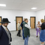 Grupa ludzi rozmawia w jasnym pomieszczeniu z szarymi kafelkami. Drzwi mają drewniane wykończenie. Z lewej strony stoi mężczyzna w kapeluszu i garniturze.