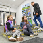 Na zdjęciu dwoje dzieci siedzi na schodach, obok przechodzi mężczyzna. Tło jest jasne, są plakaty i metalowa balustrada. Dzieci ubrane są kolorowo, a mężczyzna w kurtce.