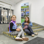 Dwie dziewczynki siedzą na schodach w budynku. Jedna ma kolorowy bezrękawnik, druga niebieską kurtkę. W tle baner promocyjny.