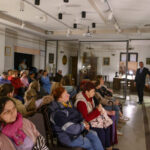 Sala z publicznością siedzącą na krzesłach; jasne światło z sufitu, biało-beżowe ściany. Prowadzący stoi obok ekranu.