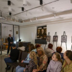 Pomieszczenie z wystawą czarno-białych fotografii na stojakach, z grupą osób siedzących na krzesłach. Ściany są w jasnych kolorach, z obrazem w złotej ramie na ścianie. Mężczyzna w garniturze przemawia do public