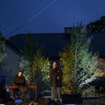 Koncert na świeżym powietrzu, wieczorem. Dwoje muzyków na scenie, wokół zielone drzewa. Niebieskie niebo w tle.