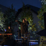 Koncert plenerowy nocą. Dwie osoby na scenie: klawiszowiec i wokalista w kapeluszu. Tło to drzewa i oświetlenie sceniczne.