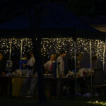 Namiot z białymi lampkami, pod którym kilka osób przygotowuje napoje. Tło jest ciemne, tworzące nastrojową atmosferę. Obok namiotu stoi stół pełen pojemników i kubków.