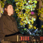 Osoba siedzi przy keyboardzie z napisem Blues Junkers. Ma na sobie ciemne ubranie i czapkę. W tle oświetlone drzewa.