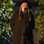 Osoba w kapeluszu i warkoczach śpiewa do mikrofonu na tle zielonych liści. Ma ciemne ubranie. Tło jest oświetlone.