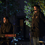 Dwóch muzyków występuje na scenie, jeden gra na klawiszach, drugi śpiewa. Noszą ciemne ubrania, w tle widać zielone liście. Światło sceniczne jest kolorowe, podkreślając atmosferę występu.