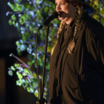 Osoba w ciemnym stroju i kapeluszu mówi do mikrofonu na tle zielonych roślin. Tło jest nieco zaciemnione. Oświetlenie skupione na osobie.