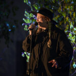 Osoba w czarnym kapeluszu śpiewa do mikrofonu. Ma dwie jasne warkocze i stoi na tle oświetlonych zielonych liści. Tło jest ciemne, nocne.